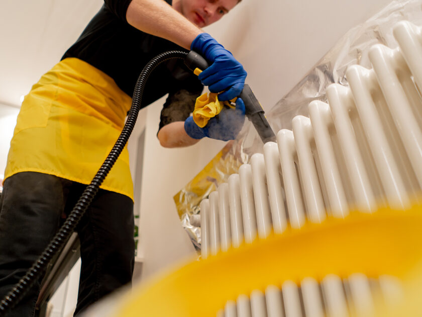 low shot in an apartment a cleaner in a yellow apron washes a white radiator with a steam generator